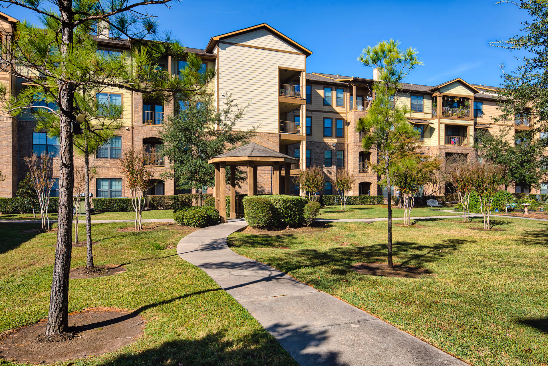 Outdoor view of a senior living facility with a paved walkway leading to a small wooden gazebo surrounded by green grass, trees, and shrubs. The background shows a multi-story building with balconies and windows under a clear blue sky.