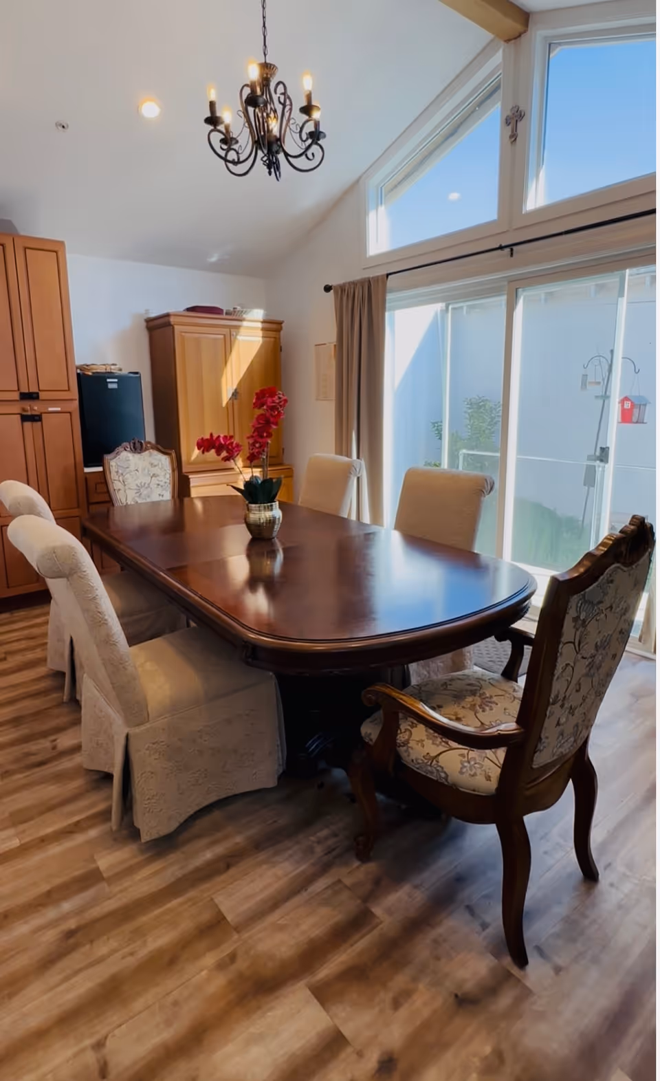 A dining room with a large wooden dining table surrounded by six upholstered chairs, including two armchairs with floral patterns. A vase with red flowers is placed on the table. The room has wooden flooring, a chandelier hanging from the ceiling, and large windows with beige curtains letting in natural light.