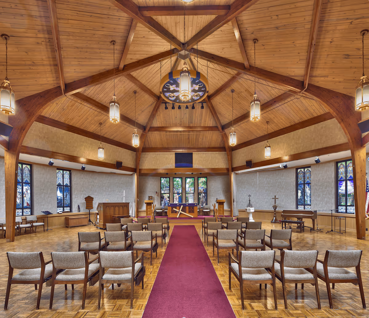 Interior view of a chapel with wooden vaulted ceiling and beams, hanging lantern-style lights, stained glass windows along the walls, rows of chairs facing an altar with religious symbols, and a red carpet aisle down the center.