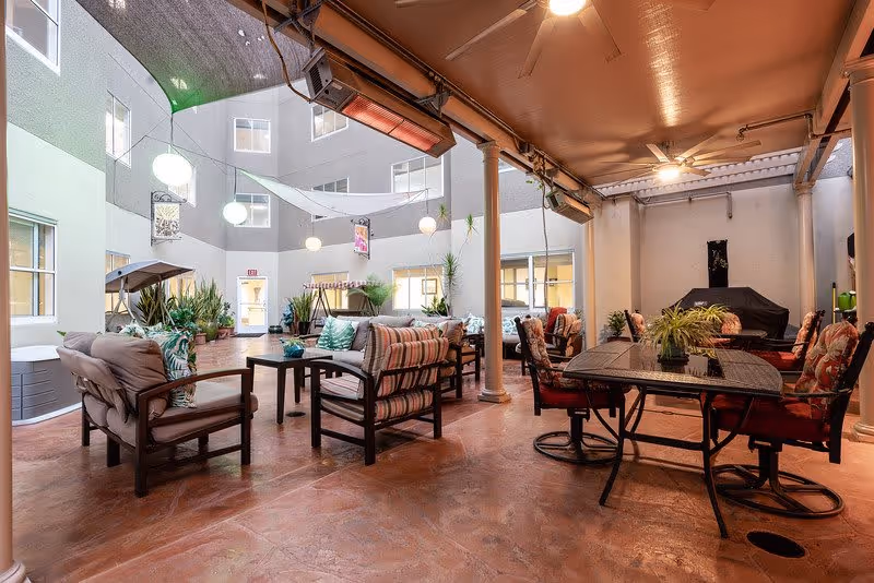 Indoor communal seating area with cushioned chairs and sofas arranged around tables, potted plants, ceiling fans, and overhead lighting, located within a multi-story building with windows overlooking the space.