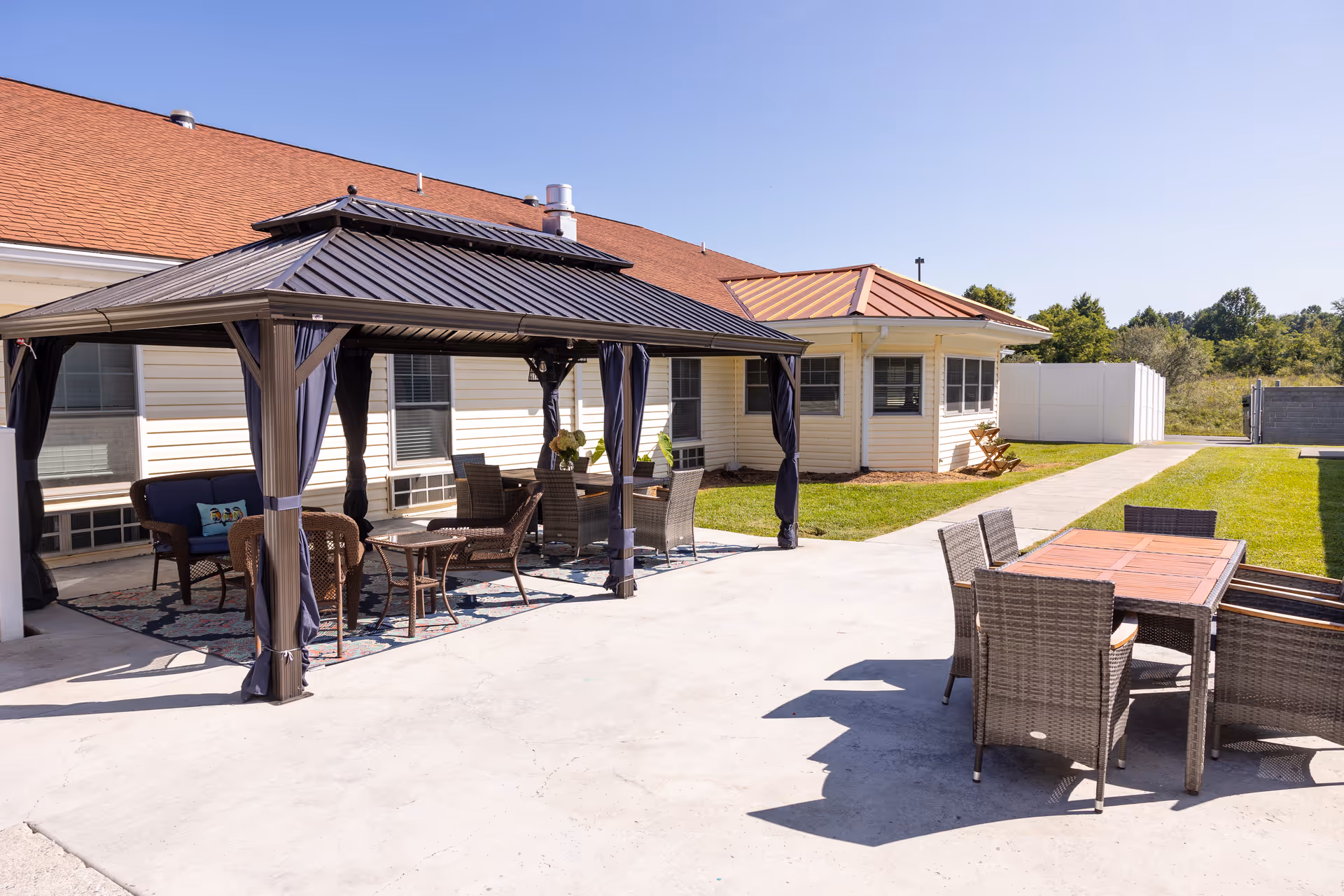 Outdoor patio area at Commonwealth Memory Care at Cedar Bluff featuring a covered gazebo with wicker chairs and a table on a patterned rug, and an uncovered dining table with six wicker chairs on a concrete surface, adjacent to a building with beige siding and red roofs under a clear blue sky.