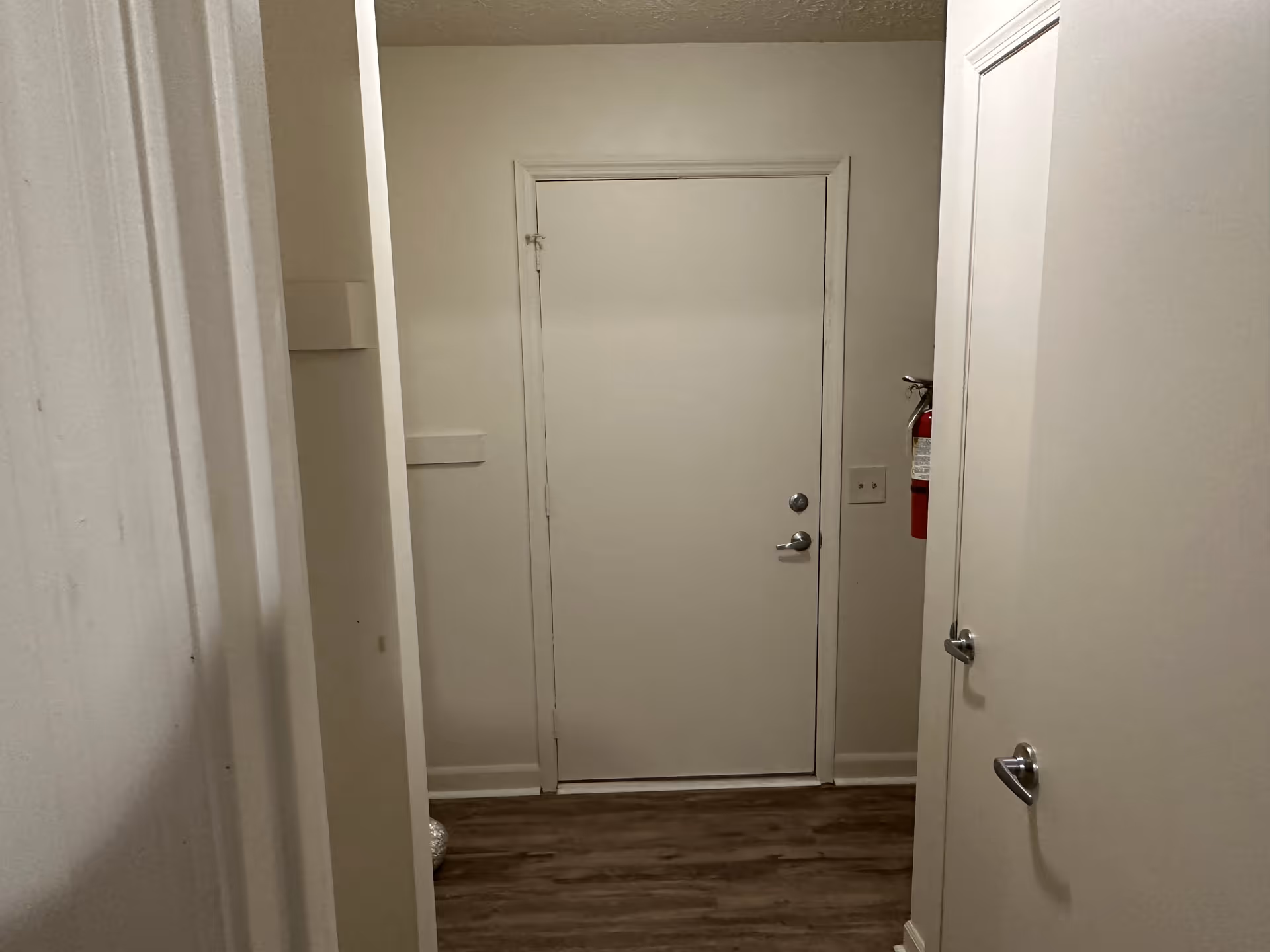 A narrow hallway with white walls and wood flooring leading to a closed white door. There is a fire extinguisher mounted on the right wall and two door handles visible on the right side, indicating doors to other rooms.