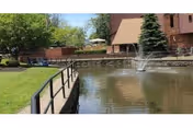 A peaceful outdoor scene at Williamstowne Senior Apartments featuring a pond with a water fountain in the center, surrounded by a black metal railing. There is green grass and trees on the left side, and a brick building with a sloped roof and a large evergreen tree on the right side.