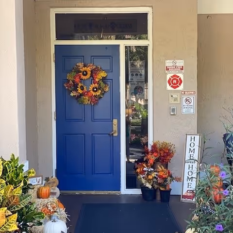Front entrance door painted blue with a fall-themed wreath hanging on it. To the right of the door is a glass panel and several signs including a fire alarm and no smoking sign. There are various potted plants and fall decorations, including pumpkins and flowers, arranged around the entrance. A vertical sign reads 'Home sweet Home'.