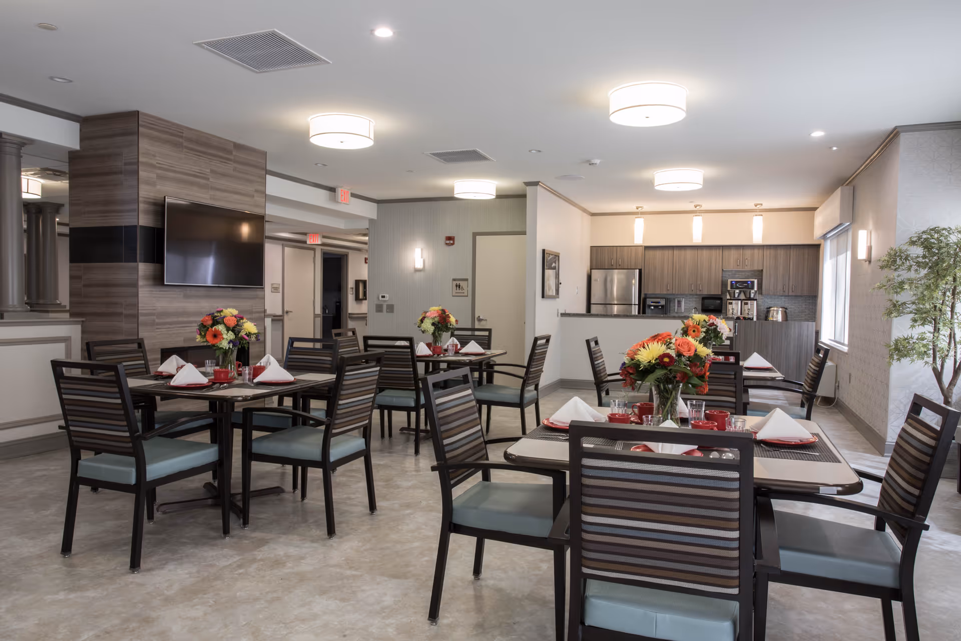 A dining room in a senior living facility with several tables set with red cups, white napkins, and floral centerpieces. The room features modern lighting, a wall-mounted TV, and a kitchenette area with stainless steel appliances in the background.