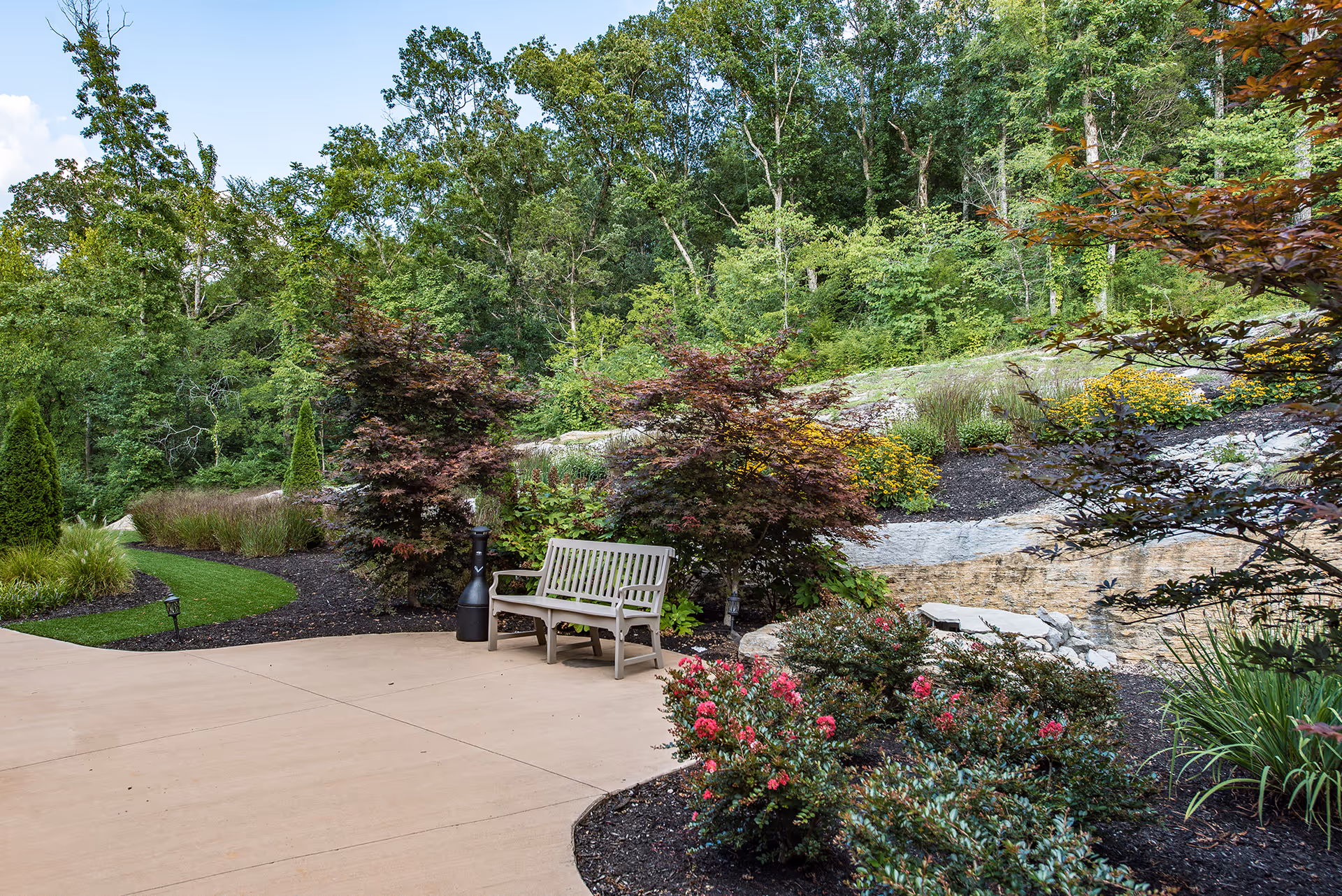 A peaceful outdoor garden area with a beige bench on a paved patio surrounded by various green shrubs, flowering plants, and trees. The background features a wooded area with dense green foliage under a clear blue sky.