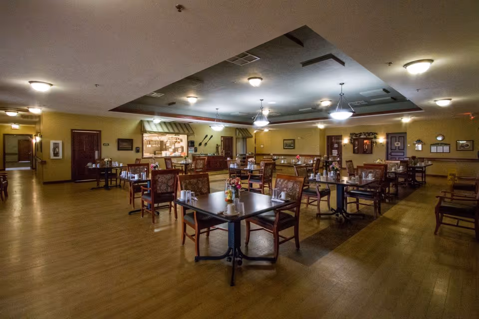 A spacious dining room with multiple tables and chairs arranged neatly. Each table has place settings including cups and small flower arrangements. The room has warm lighting with ceiling fixtures and a serving window area in the back wall. The floor is wooden, and the walls are painted a soft yellow color.
