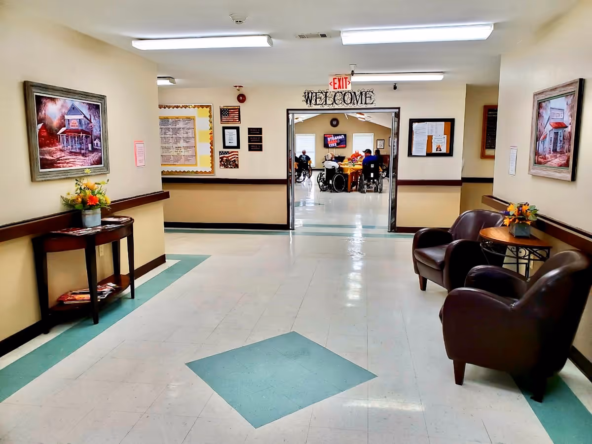 A clean and well-lit hallway in a nursing and rehabilitation facility with beige walls and a tiled floor featuring green diamond patterns. On the left side, there is a small table with a flower arrangement and magazines underneath, and a framed painting above it. On the right side, there are two brown leather chairs with a small round table between them, also with a flower arrangement. At the end of the hallway, double doors lead to a common room where several people in wheelchairs are gathered around a table. Above the doors, a decorative sign reads 'WELCOME' and an exit sign is visible.