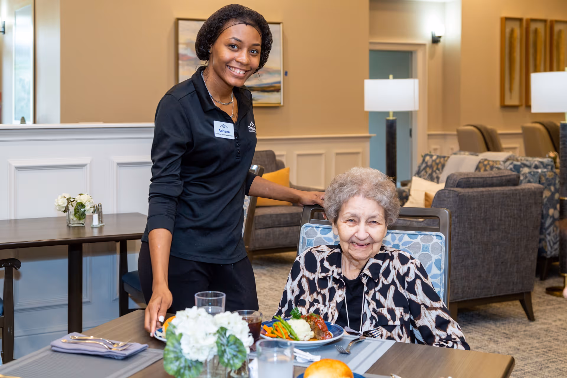 A smiling caregiver stands beside an elderly woman seated at a dining table with a plate of food in a bright communal dining area.