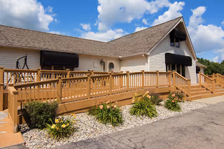 Exterior view of a single-story building with beige siding and a brown shingled roof under a blue sky with scattered clouds. The building features black awnings over the windows and a wooden ramp with railings leading to the entrance. There are small landscaped areas with green shrubs and flowering plants along the front of the building.