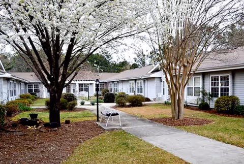 Outdoor courtyard area of a senior living facility with a concrete walkway, blooming trees, shrubs, and single-story buildings with multiple windows surrounding the courtyard.