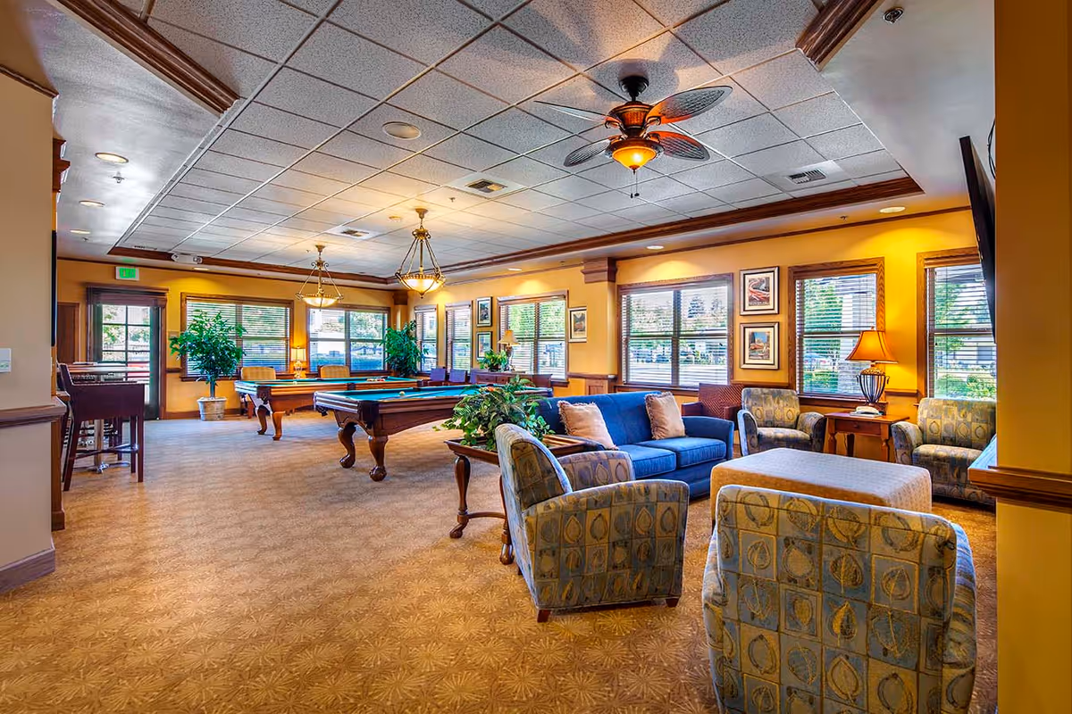 A spacious and well-lit common area in a senior living facility featuring a blue sofa, patterned armchairs, a large ottoman, two pool tables, ceiling fans, large windows with blinds, framed artwork on the walls, and potted plants.