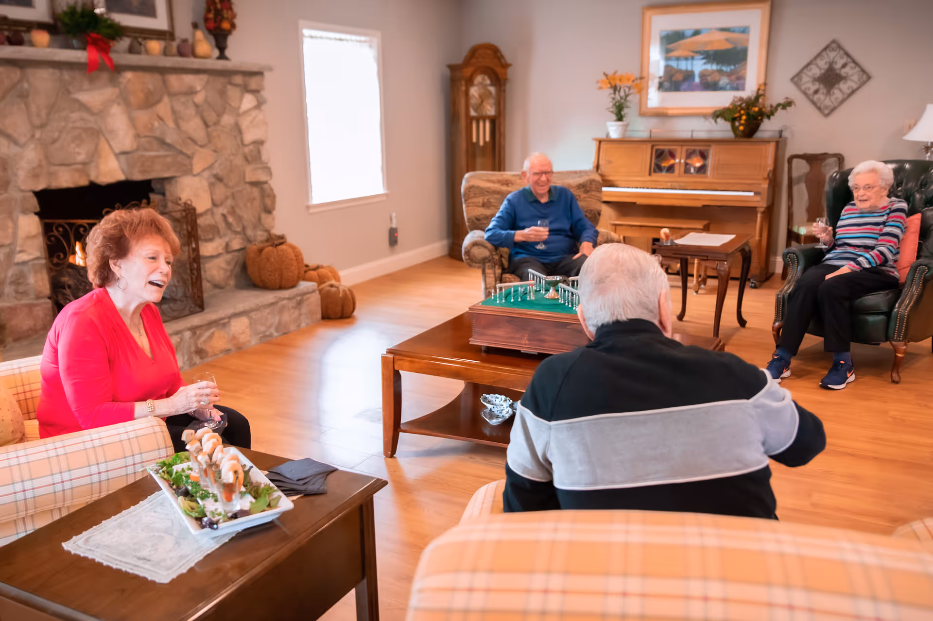 A cozy senior living room with four elderly people sitting and enjoying drinks. The room features a stone fireplace with a fire burning, a wooden coffee table with a game on it, a piano against the wall, and autumn decorations including pumpkins. The atmosphere is warm and inviting.