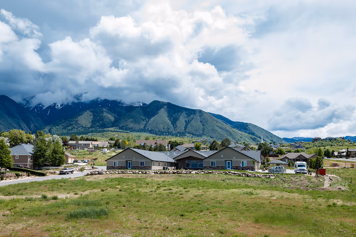 A single-story building with a gray roof and beige walls situated in a green grassy area with a mountainous backdrop under a partly cloudy sky. Several houses and vehicles are visible around the building.
