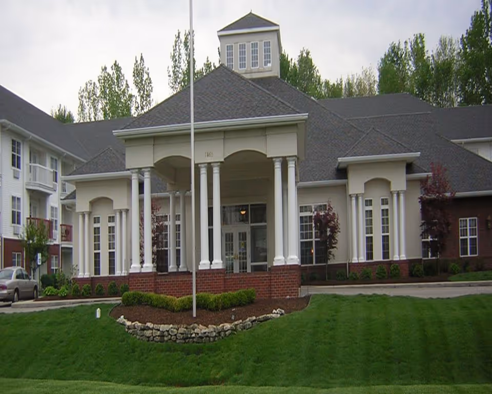 Front exterior view of Wyndmoor Assisted Living facility showing a large building with a covered entrance supported by white columns, manicured green lawn, and trees in the background under a cloudy sky.