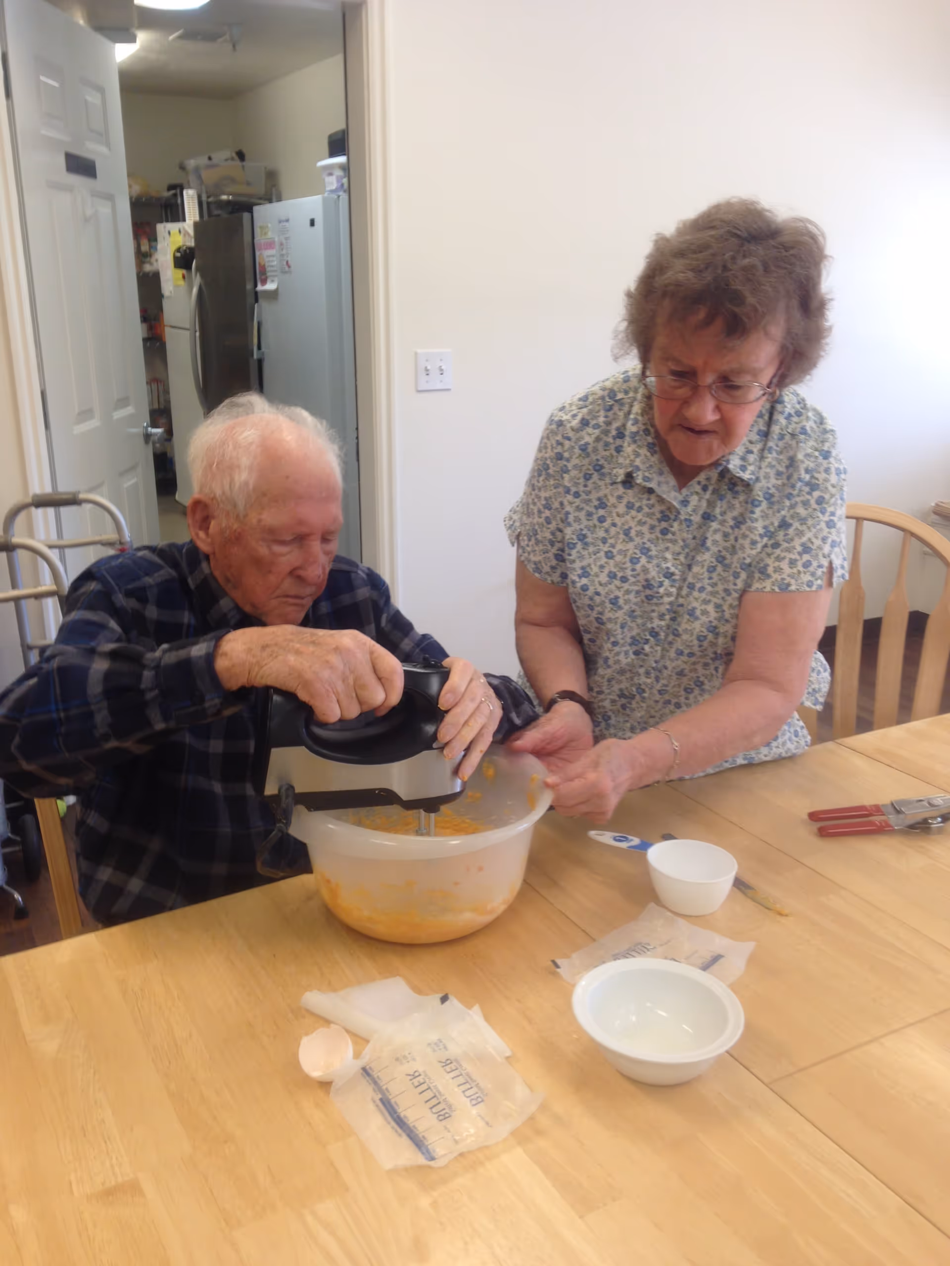 An elderly man and woman are sitting at a wooden table in a kitchen. The man is using a hand mixer to blend ingredients in a large plastic bowl, while the woman watches and assists. On the table, there are empty bowls, an eggshell, and some packaging. In the background, a refrigerator and pantry shelves are visible through an open door.