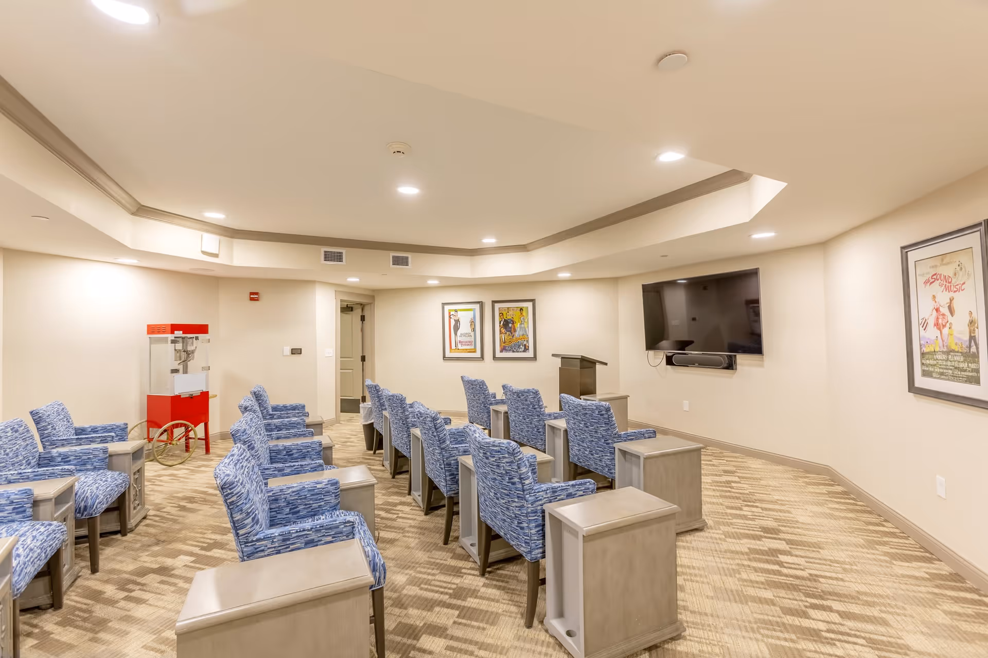 A small theater-style room with rows of blue patterned chairs facing a wall-mounted flat screen TV and a podium. The room has beige walls, carpeted floor, framed posters on the walls, and a red popcorn machine in the corner.