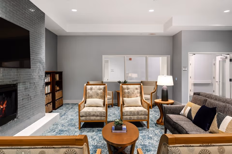 Well-lit communal living room with upholstered armchairs and a sofa arranged around a coffee table, a fireplace and a wall-mounted TV.