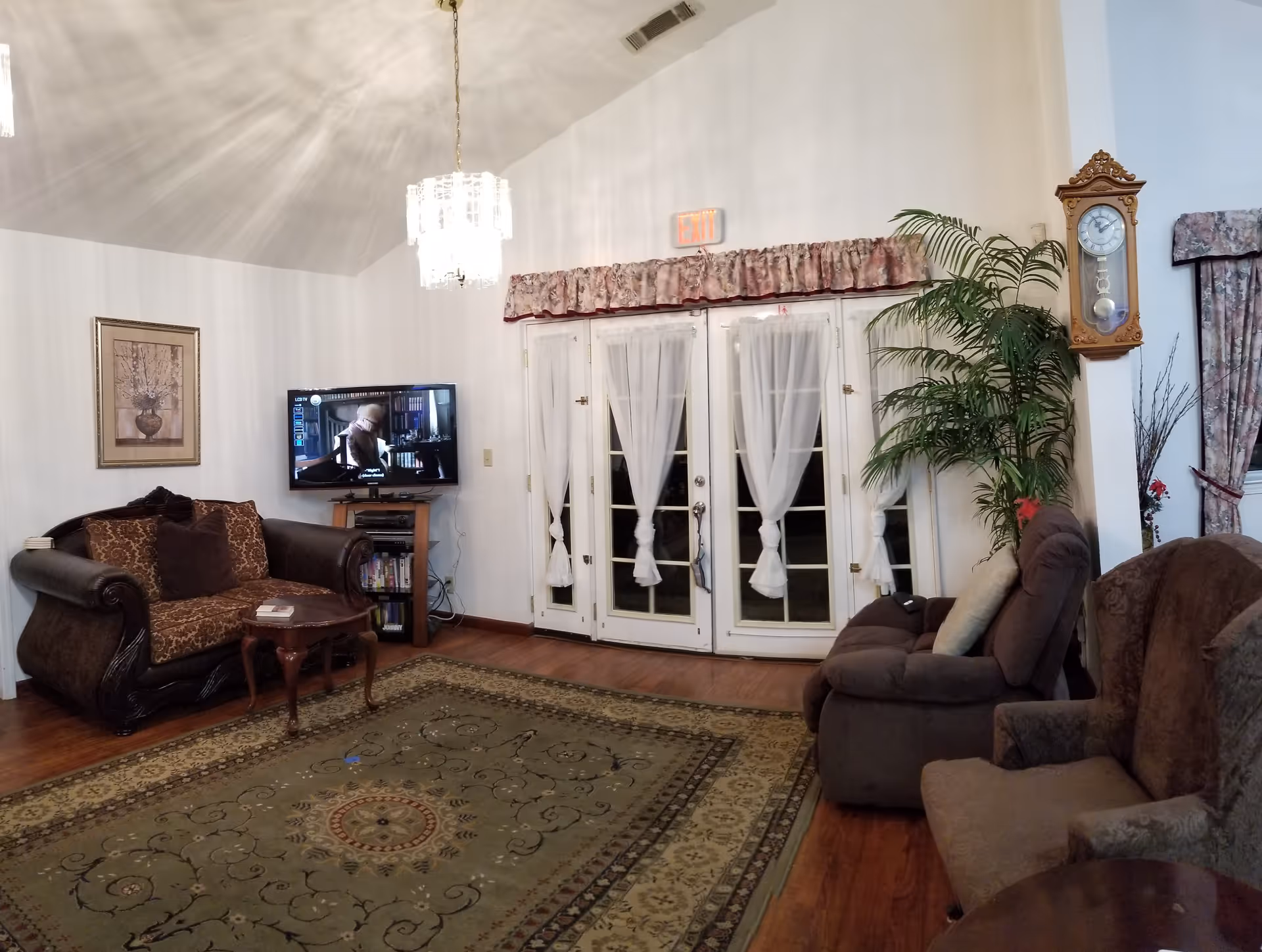 A cozy living room with a patterned area rug, a dark brown ornate loveseat with cushions, a small wooden coffee table, a wall-mounted TV showing a scene from a movie, a tall green potted plant, a wooden wall clock, and French doors with sheer white curtains and floral valances. The room has hardwood floors and a chandelier hanging from the ceiling.