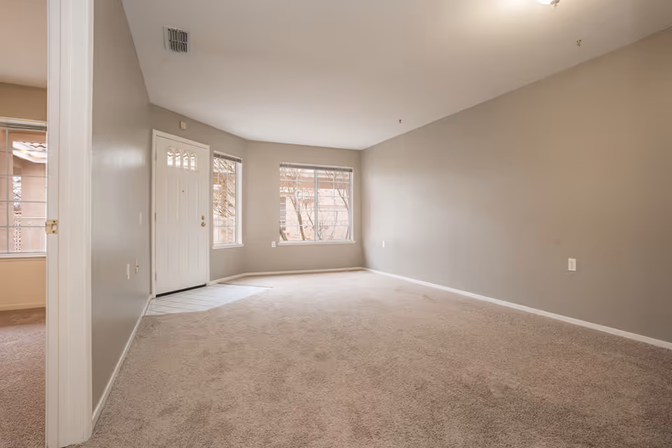 Empty living room area with beige carpet and light gray walls, featuring a white front door with windows on either side and a window on the adjacent wall letting in natural light.