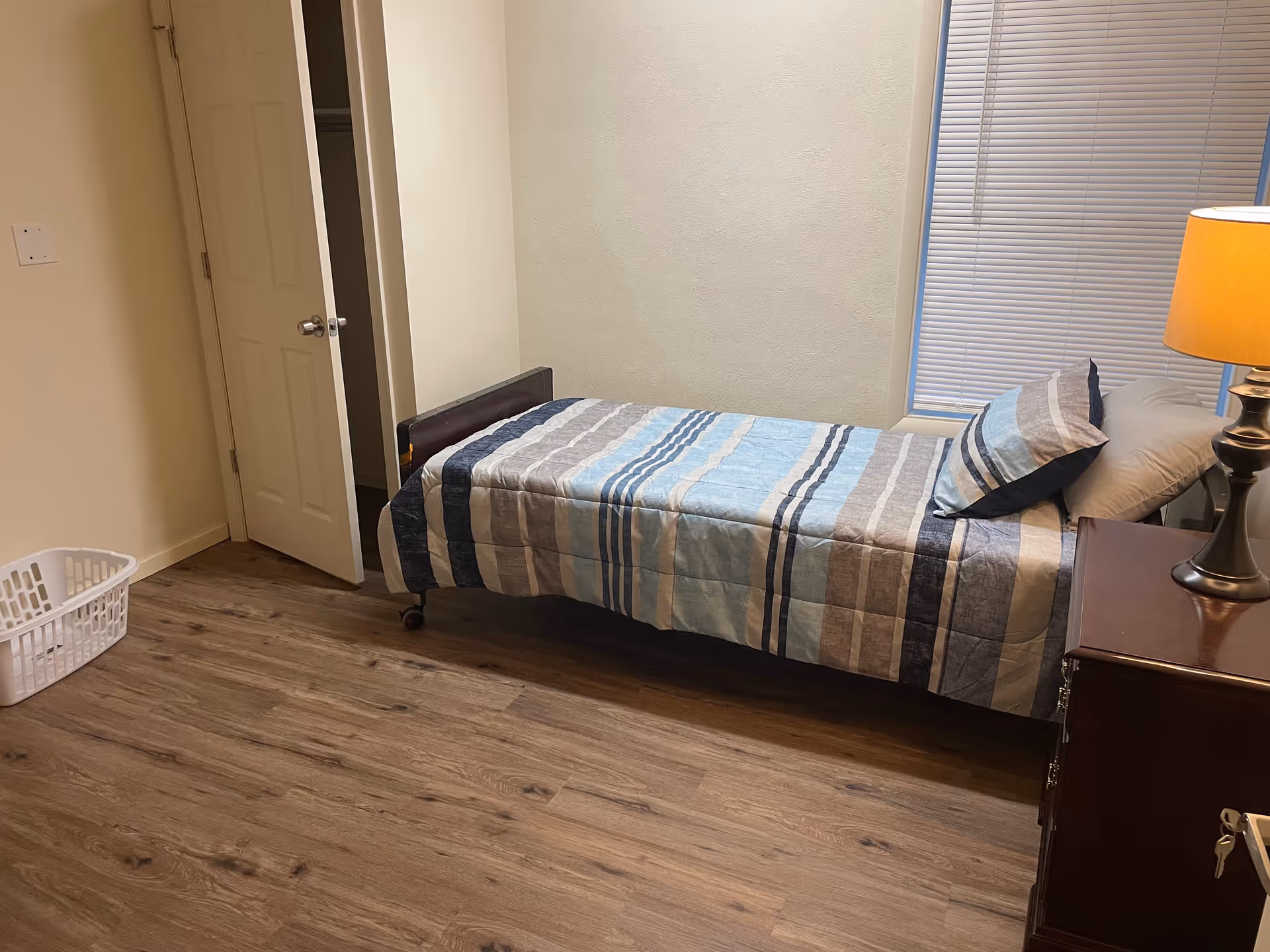 A small bedroom with a single bed covered in a striped blue, gray, and white comforter. There is a matching pillow on the bed. Next to the bed is a wooden nightstand with a lamp on it. A window with closed blinds is behind the bed. The floor is wood, and there is a white laundry basket on the floor near a partially open door.