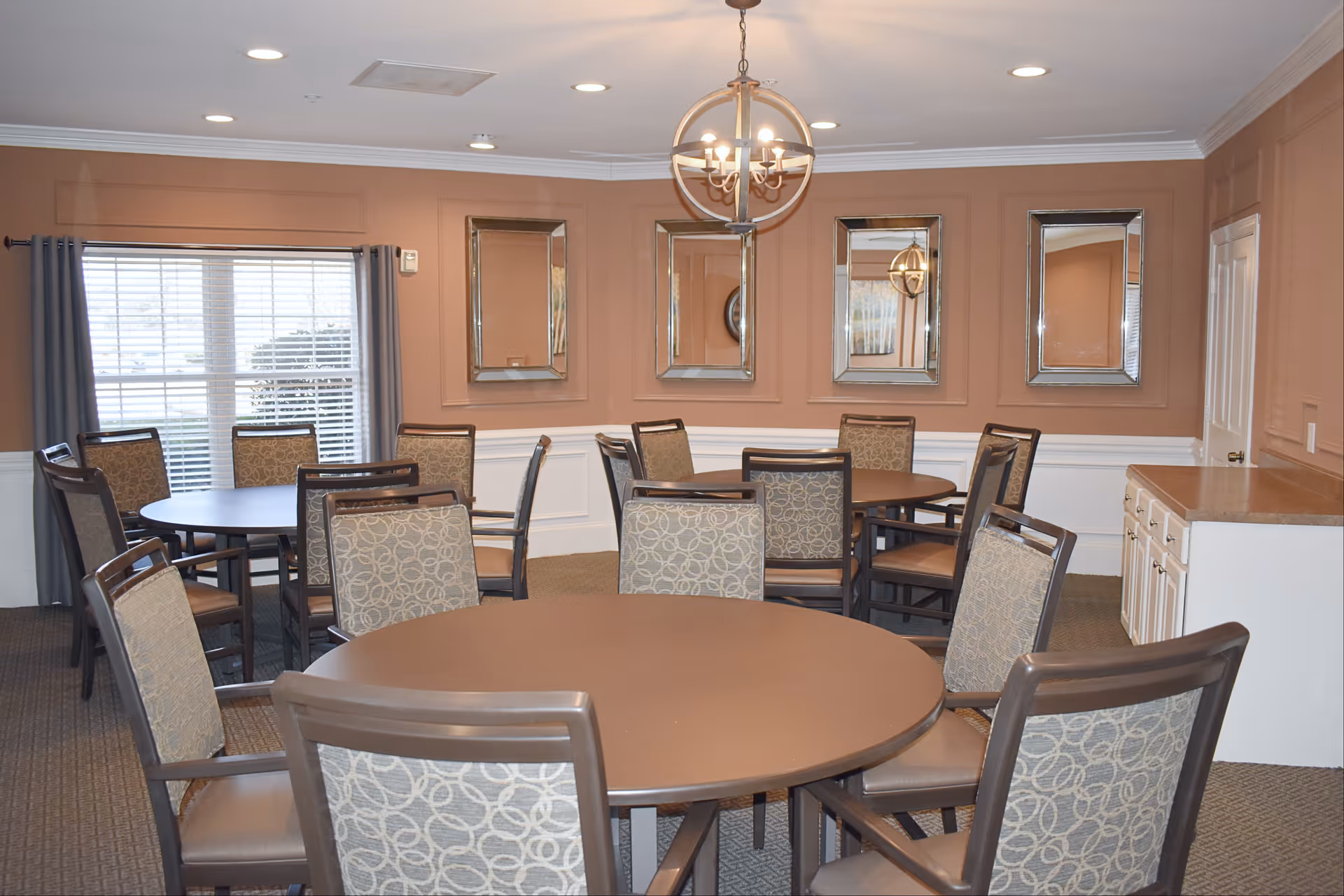 A dining room with multiple round tables and chairs arranged around them. The walls are painted a warm brown color with white wainscoting. There are four rectangular mirrors hanging on the wall and a window with gray curtains letting in natural light. A chandelier with multiple lights hangs from the ceiling. There is also a white cabinet with a countertop on the right side of the room.