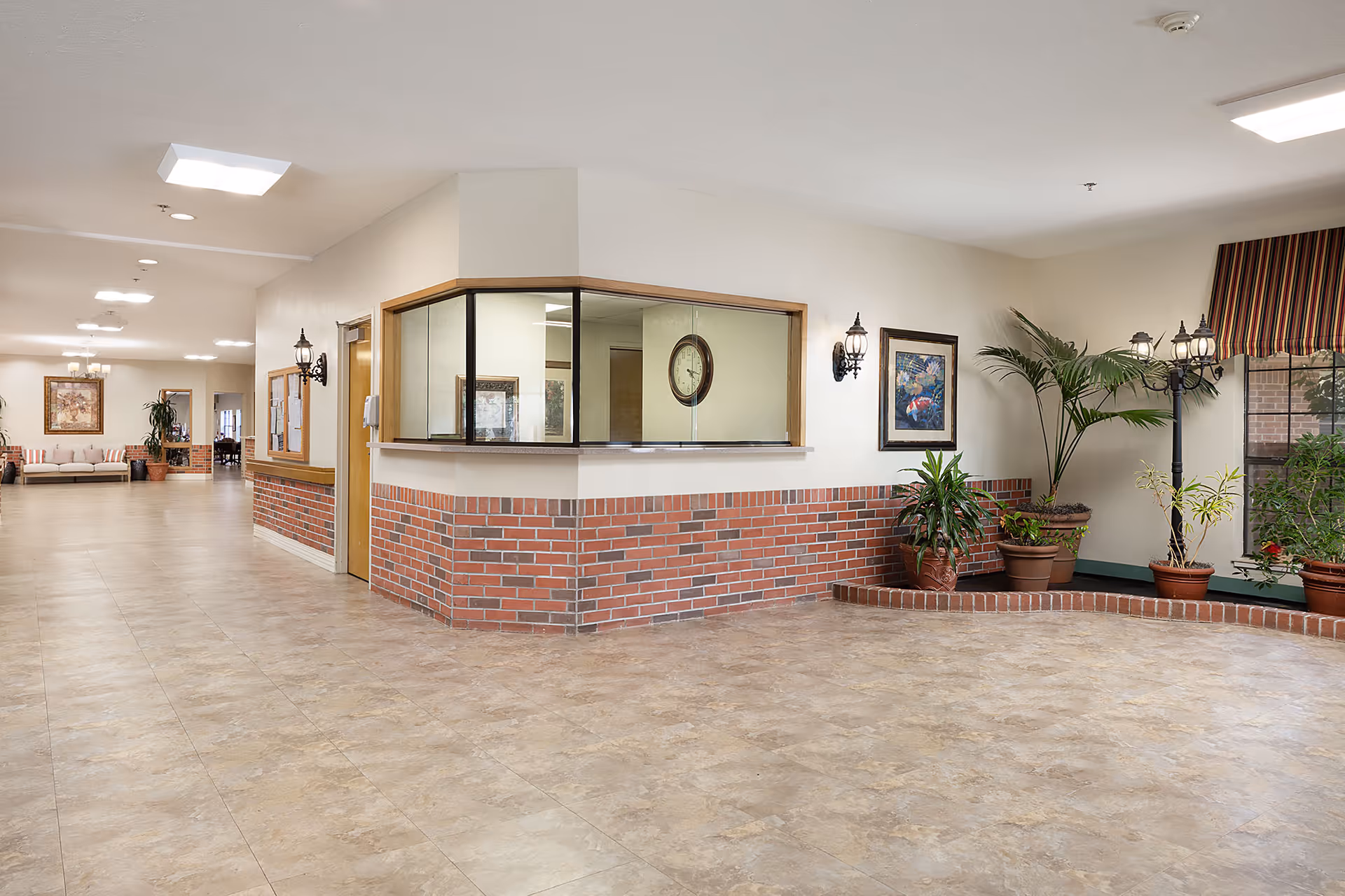 Spacious nursing facility lobby with a reception window set in a brick half-wall, potted plants, seating area down the hallway, and tiled floors.