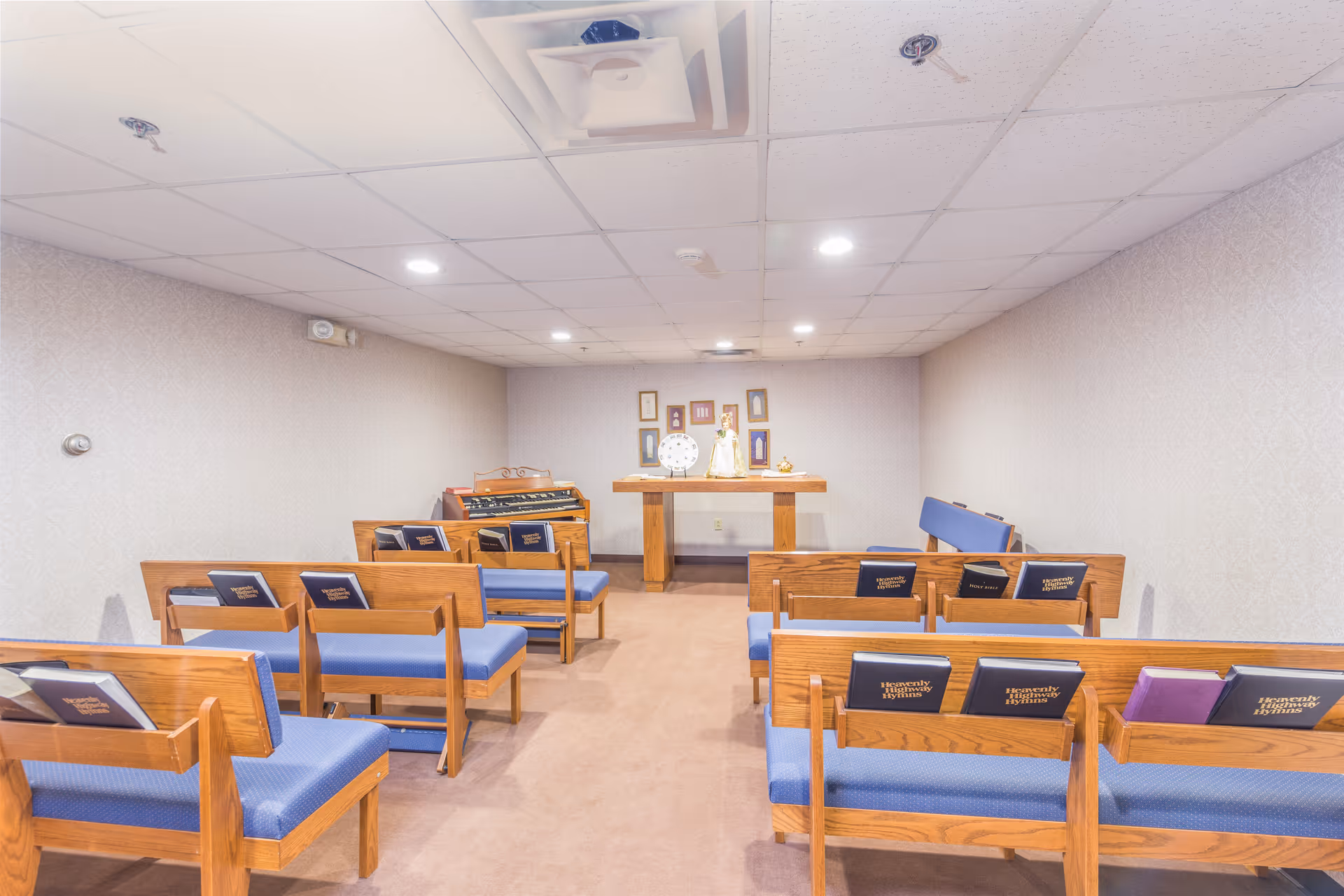 A small chapel or prayer room with wooden pews featuring blue cushions and hymnals placed on the backrests. At the front, there is a wooden altar table with religious items and framed pictures on the wall behind it. The room has a carpeted floor and a drop ceiling with recessed lighting.