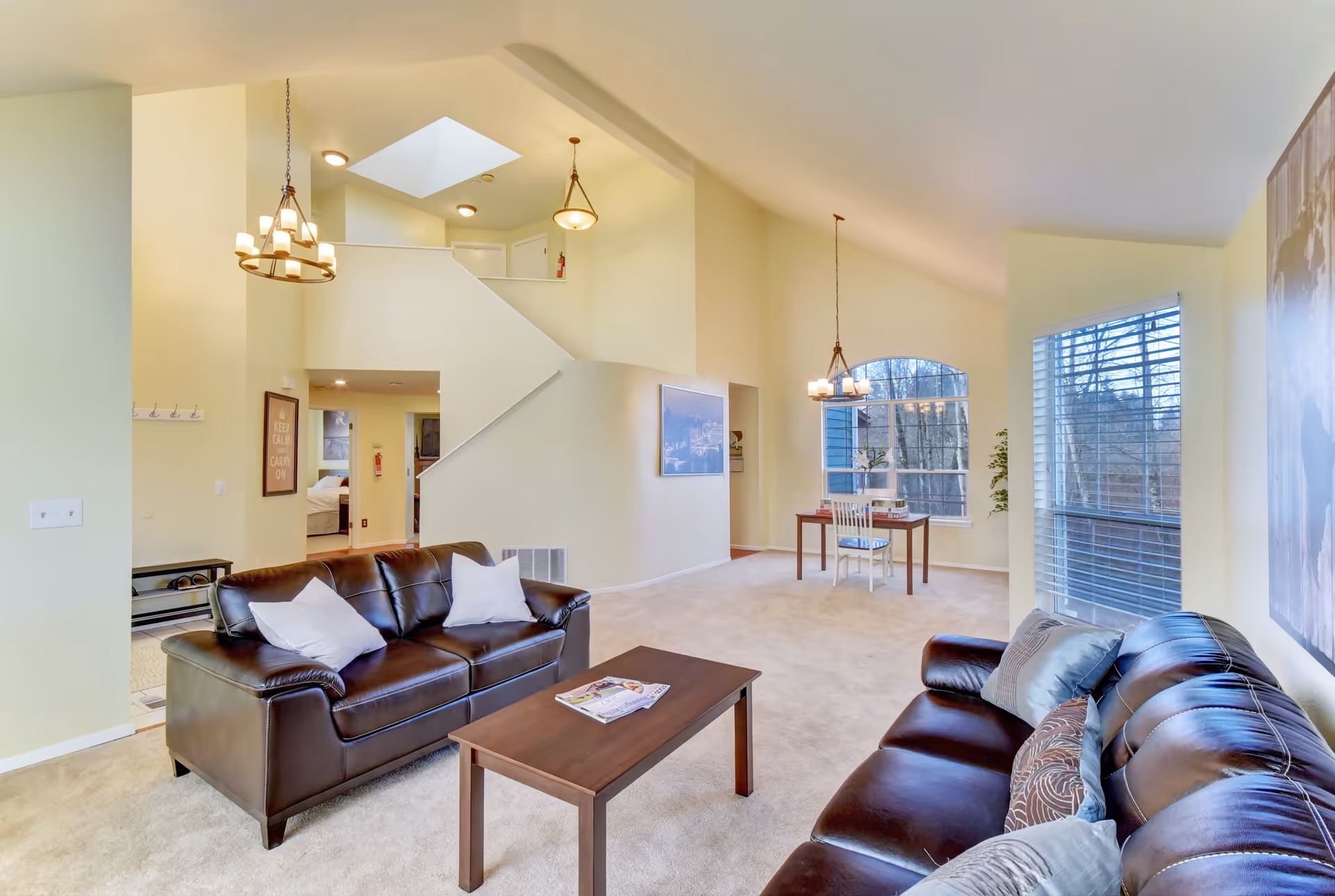 A bright and spacious living room with two dark brown leather sofas facing each other, a wooden coffee table in between, and a small dining table with two chairs near large windows. The room has beige walls, carpeted floor, and a staircase leading to an upper level. There are pendant lights hanging from the ceiling and a skylight above the staircase.