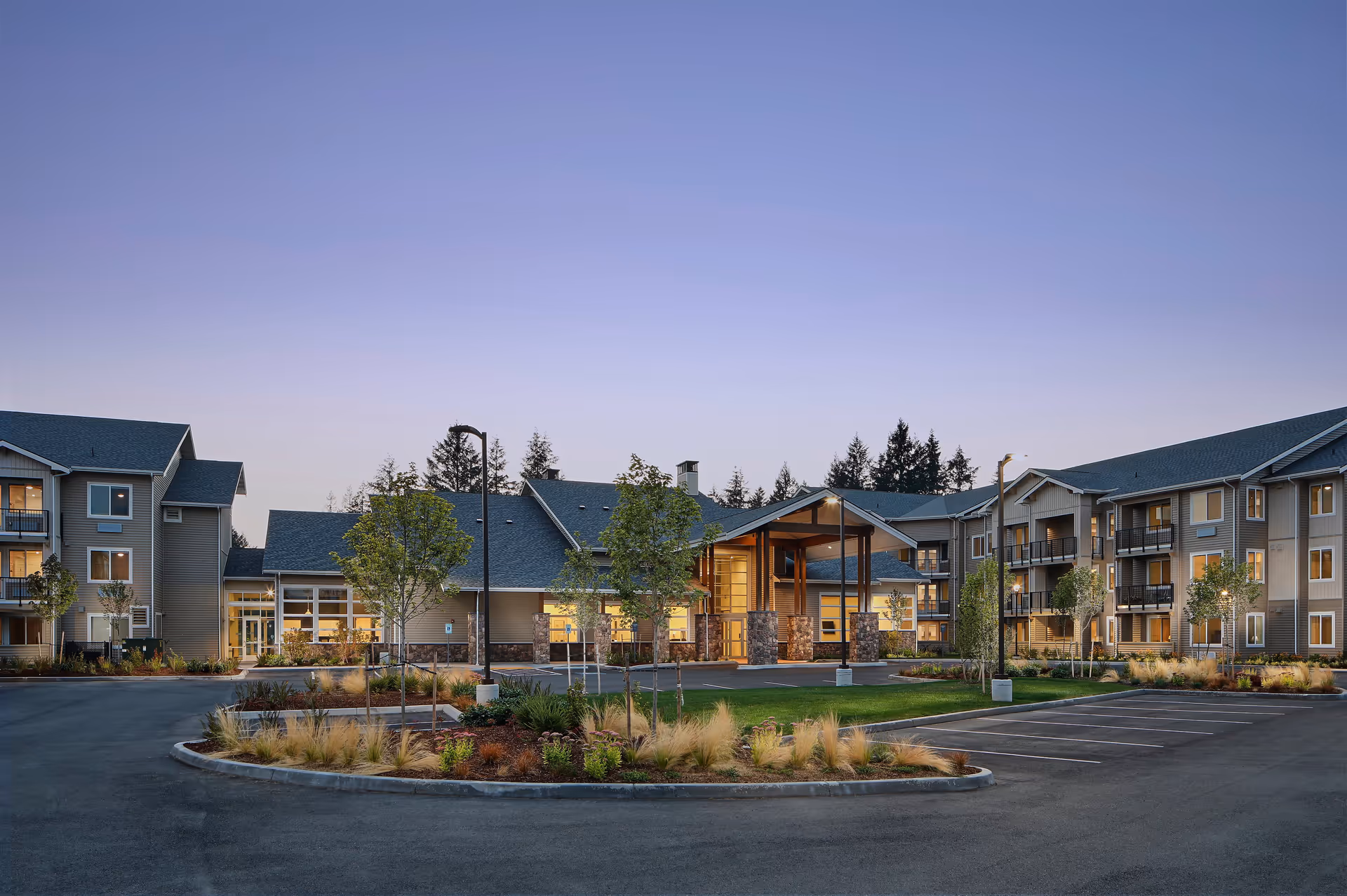 Exterior view of Revel Lacey senior living facility at dusk, showing a large building with multiple floors, balconies, a covered entrance, landscaped greenery, and an empty parking lot.
