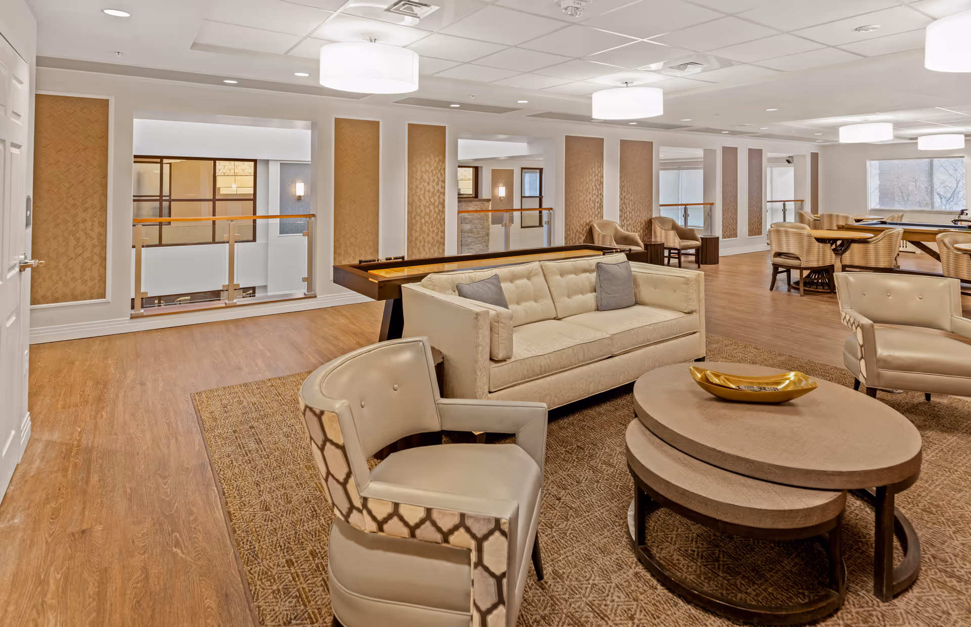 A spacious and well-lit common area in a senior living facility featuring a beige sofa with two gray pillows, two beige armchairs with patterned backs, a set of nested round coffee tables with a decorative golden bowl, a shuffleboard table, and several wicker chairs around tables near large windows.