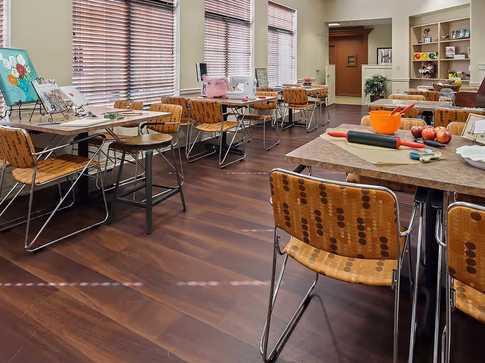 Communal activity/dining room with multiple tables and patterned chairs, craft supplies and kitchen items on the tables, and large windows with blinds.