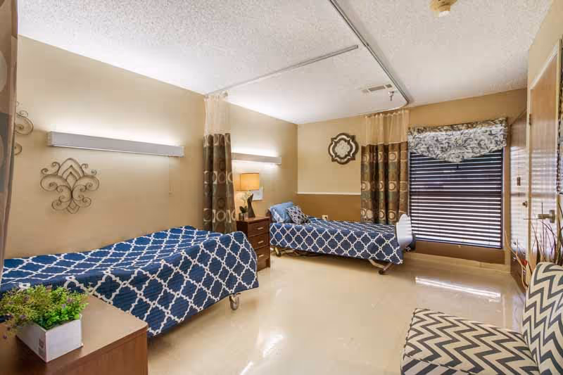 Shared nursing facility bedroom with two single beds, a nightstand, patterned bedspreads and a window with blinds.