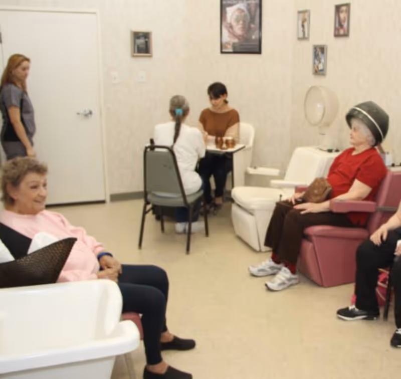A room in a senior living facility where elderly women are seated. One woman is sitting under a hair dryer, another is seated on a chair with a basket on her lap, and two women are sitting at a small table engaged in an activity. A staff member stands near a closed door. The room has light-colored walls with framed pictures.