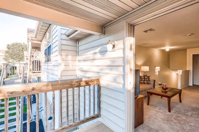 View of a small outdoor balcony with wooden railings attached to a senior living apartment. The balcony overlooks a grassy area and neighboring buildings. The sliding glass door is open, showing a living room inside with a beige carpet, a wooden coffee table with a red candle, a couch with patterned cushions, a lamp on a side table, and a hallway leading to other rooms.