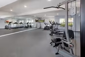 A fitness room with various exercise equipment including treadmills, stationary bikes, and weight machines. The room has large mirrors on one wall, windows letting in natural light, and a ceiling fan.