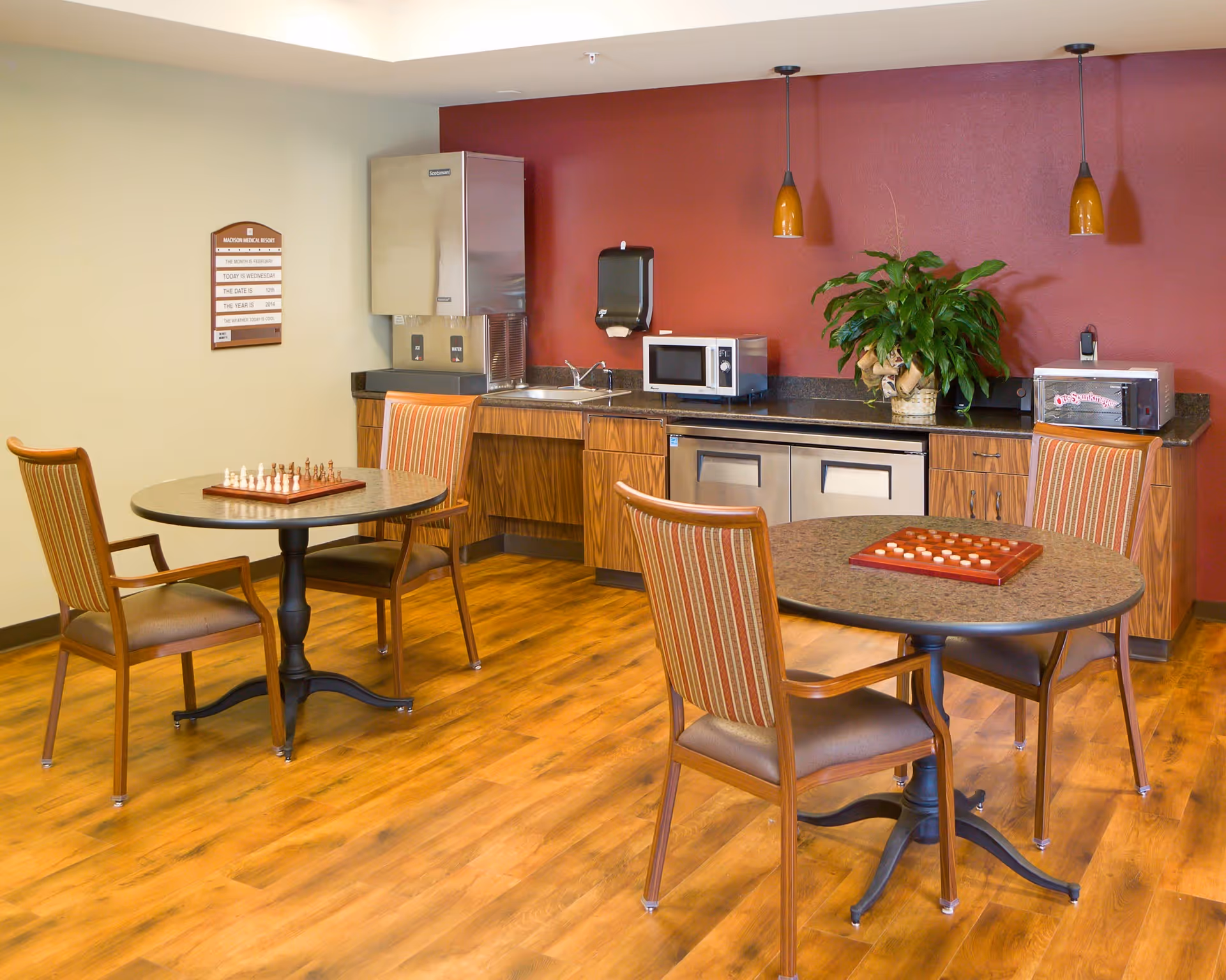 Small common room with two round game tables and chairs in front of a kitchenette with a sink, microwave, and cabinetry against a red wall.