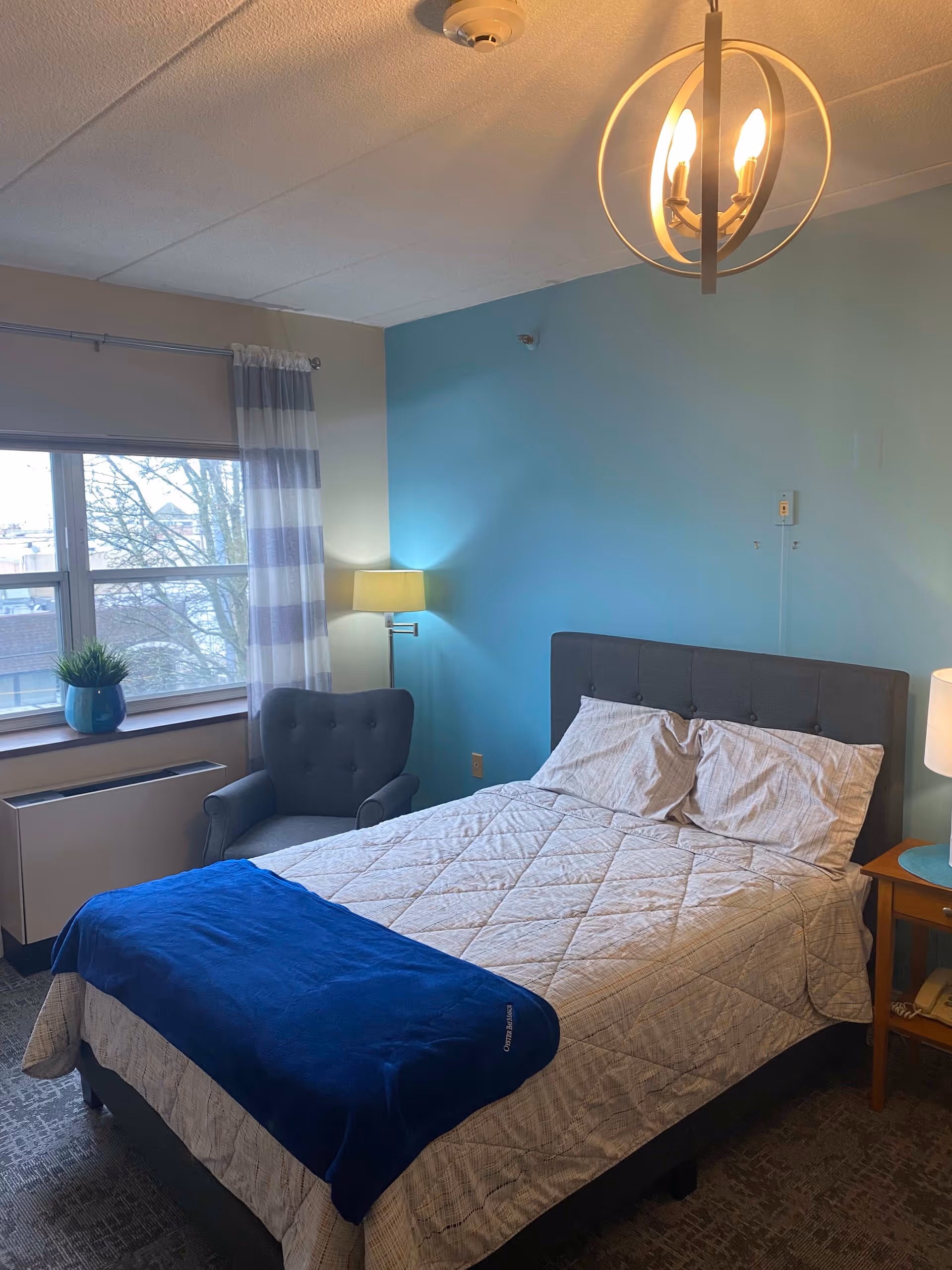 A cozy bedroom with a neatly made bed featuring white and beige bedding and a blue throw blanket. There is a gray upholstered headboard, a gray armchair next to a window with striped curtains, a small wooden nightstand with a lamp, and a ceiling light fixture. The walls are painted light blue and beige.
