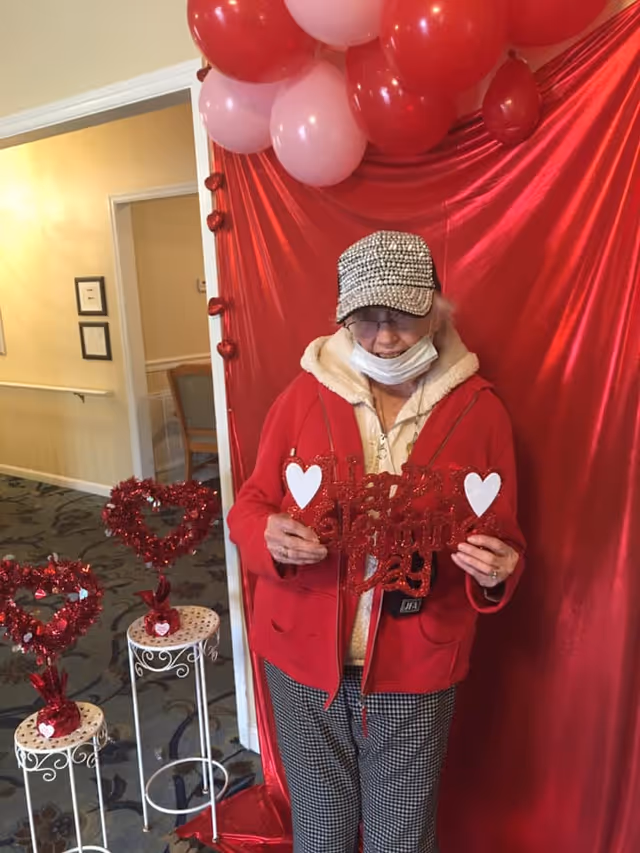 An elderly woman wearing a red jacket, checkered pants, a rhinestone-studded cap, and a face mask pulled down under her chin stands in front of a red backdrop decorated with red and pink balloons. She is holding a glittery red sign that says 'Happy Valentine's Day' with white heart decorations. There are two small white tables with red heart-shaped decorations on them to her left.