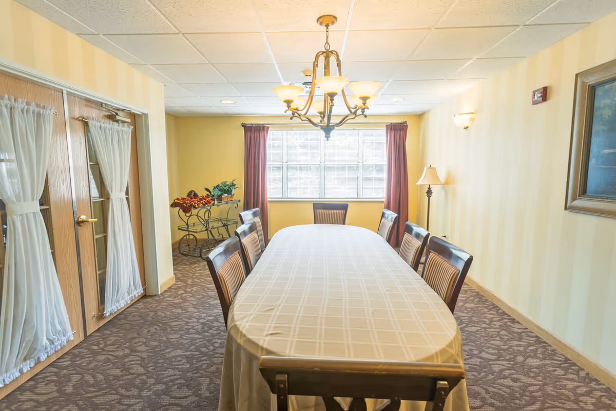 A bright dining room with a long table covered by a tablecloth, chairs around it, a chandelier above and a window at the far end.