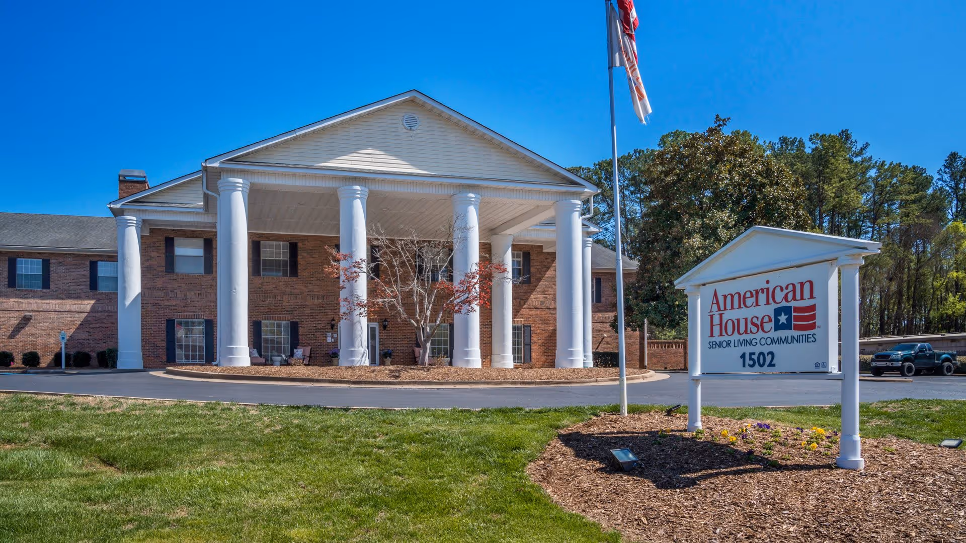 Front exterior view of American House Hamilton Place senior living community building with large white columns, a flagpole with an American flag, and a sign displaying the facility name and address 1502. The building is made of brick and has multiple windows with black shutters. There is a green lawn and some landscaping in front of the building.