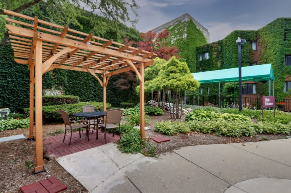 Outdoor garden area at PA Peterson at the Citadel featuring a wooden pergola with a table and four chairs underneath. The surrounding area includes various green plants, bushes, and trees. The building in the background is covered with green ivy, and there is a green canopy over a walkway with a sign indicating the main entrance.