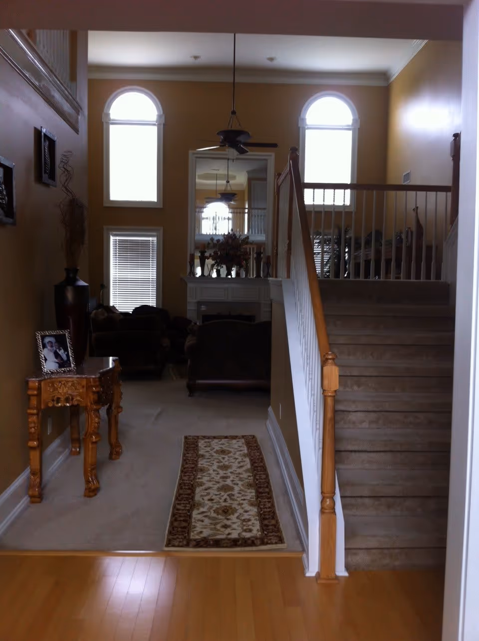 Entry hallway leading into a two-story living room with a staircase on the right, hardwood floors, arched windows, and a fireplace.