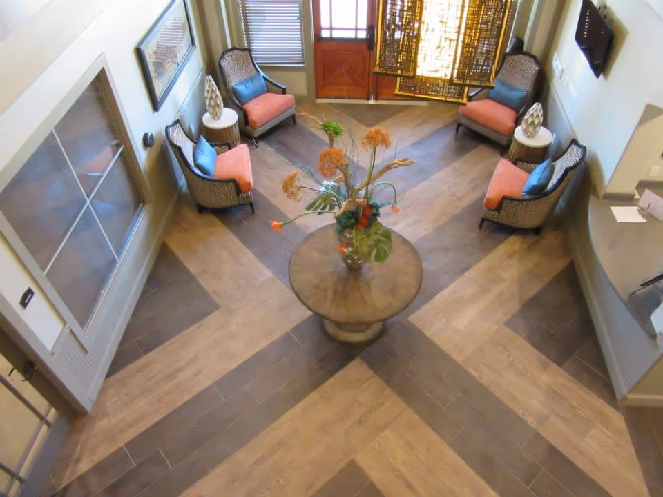 A cozy seating area with four cushioned armchairs arranged around a round wooden table with a floral centerpiece. The floor has a patterned wood design, and there is a door with windows and a decorative screen in the background. The space appears well-lit and inviting.