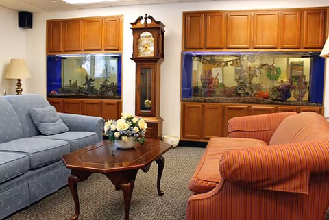 A cozy living room with a blue patterned sofa and a red striped sofa facing each other across a wooden coffee table with a flower arrangement. Behind the sofas, there are two large built-in aquariums set in wooden cabinetry and a tall grandfather clock between them. A floor lamp is visible next to the blue sofa.