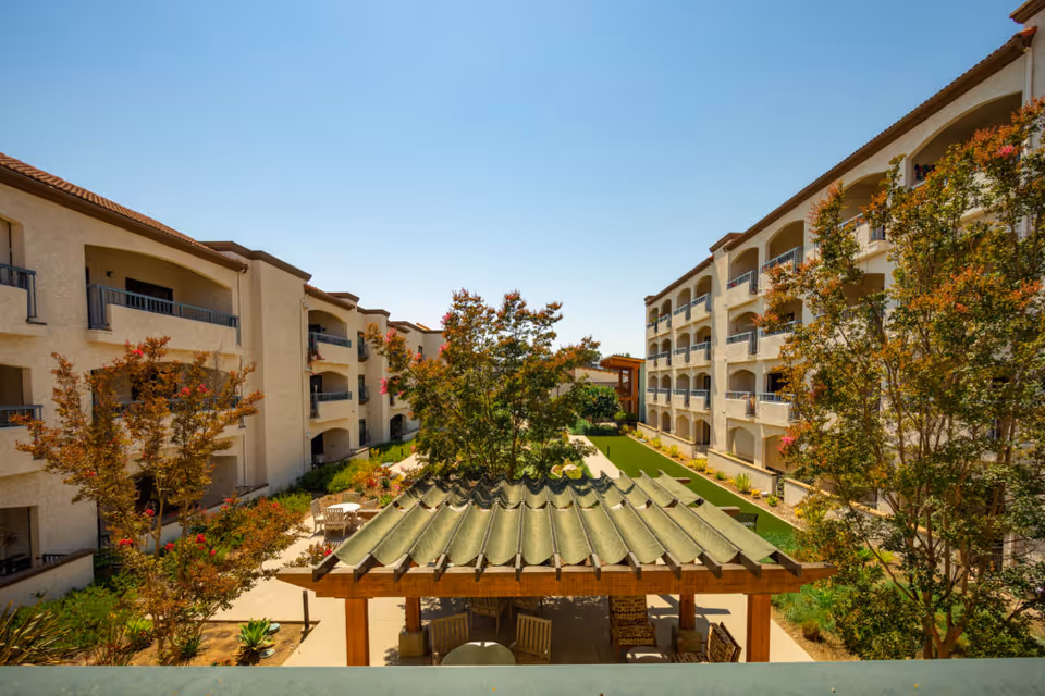 Outdoor courtyard area of Casa de las Campanas featuring a covered seating area with wooden chairs, surrounded by trees and plants, with multi-story residential buildings on both sides under a clear blue sky.