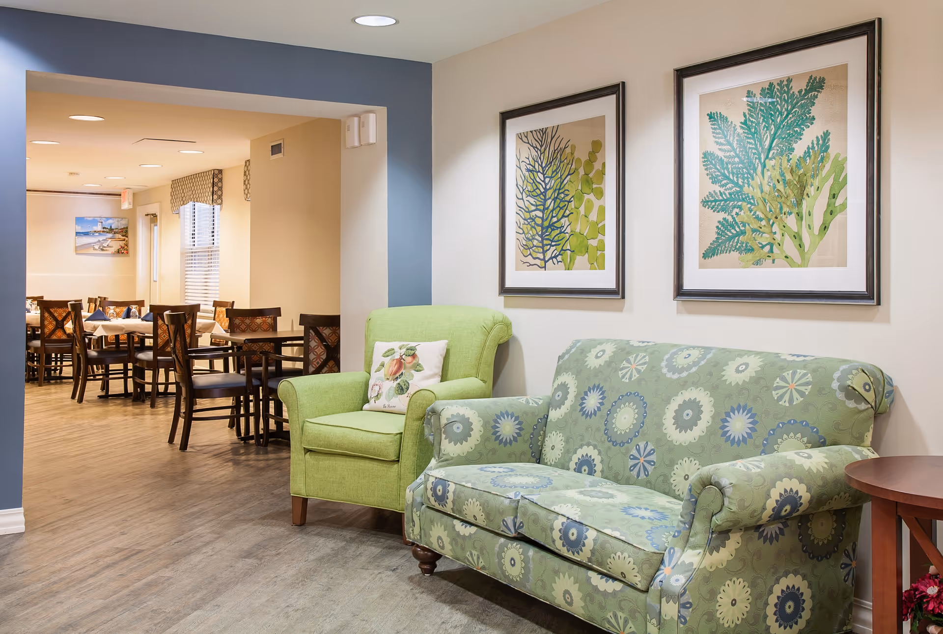 A cozy seating area with a green patterned sofa and a light green armchair with a decorative pillow, two framed botanical prints on the wall above, and a view into a dining area with multiple tables and chairs in the background.