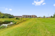 Wide grassy field with a small pond on the left side and a large multi-story residential building in the distance under a blue sky with a few clouds.