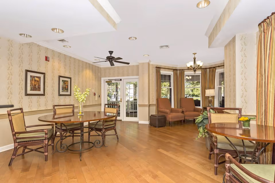 Sunlit common room with round wooden dining tables and chairs, upholstered lounge chairs by bay windows, hardwood floors, and patterned wallpaper.