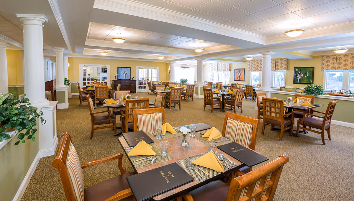 A spacious and well-lit dining room with multiple wooden tables and chairs arranged neatly. Each table is set with yellow folded napkins, glasses, silverware, and menus. The room features beige carpeting, light yellow walls, white columns, and several windows allowing natural light. Decorative paintings and plants add to the welcoming atmosphere.